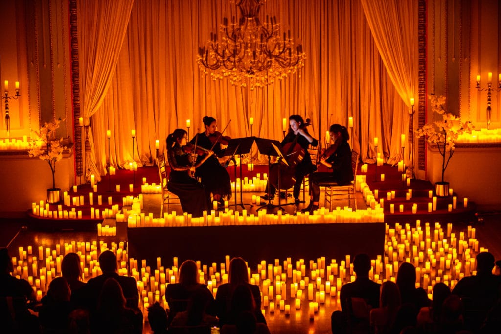 a group of string musicians perform on stage surrounded by hundreds of candles as audience watches