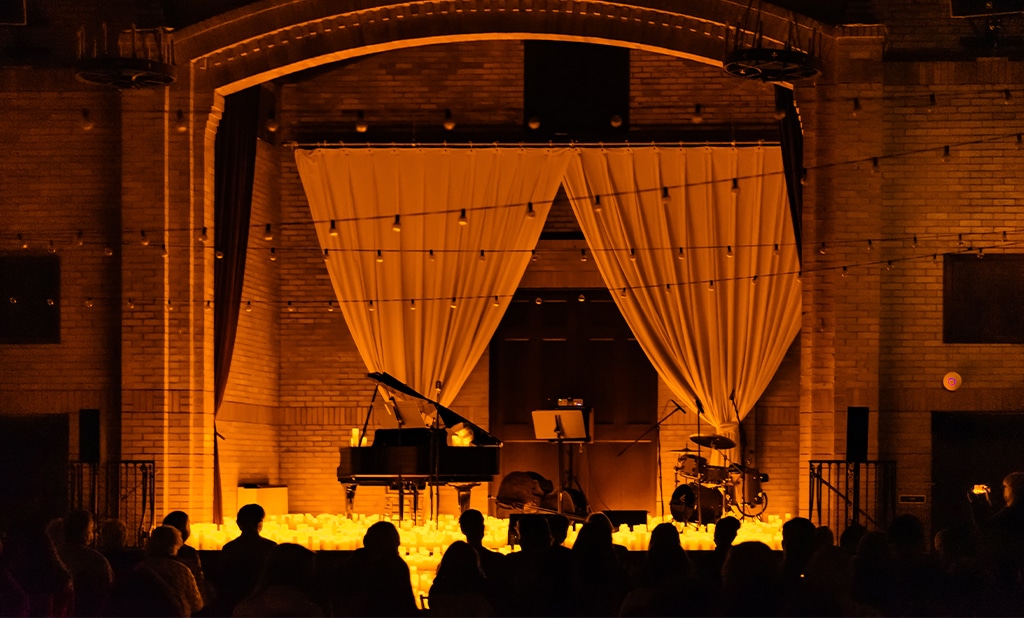 A silhouette of seated audience members across a candlelit stage with a grand piano at St Francis Hall