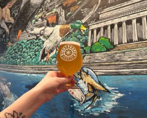A person holds a pint of beer up against a mural of swans on a lake.